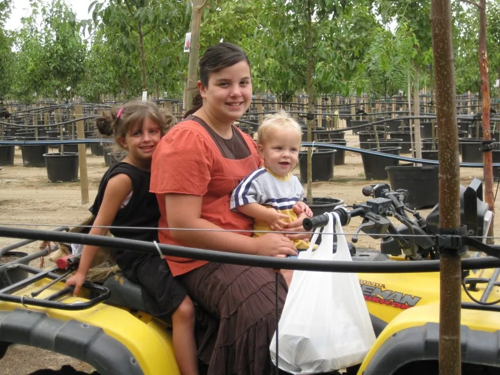 Three children posing on a yellow ATV among rows of potted trees