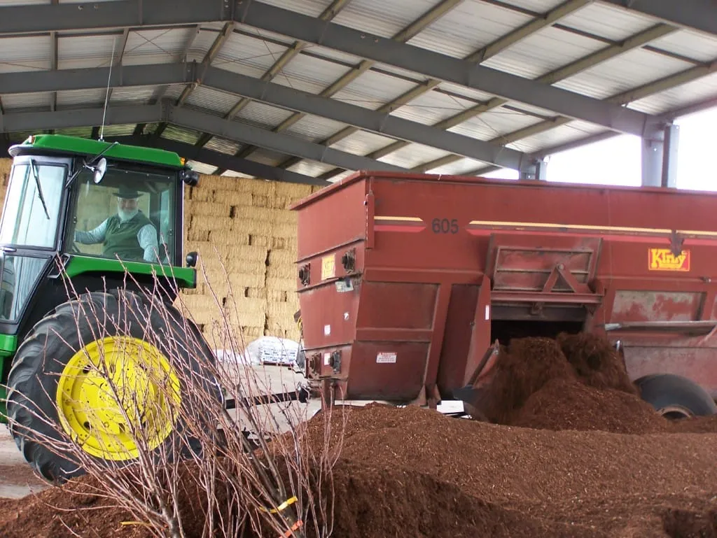 Man driving a John Deere tractor dumping mulch from a red Kirby wagon under a barn
