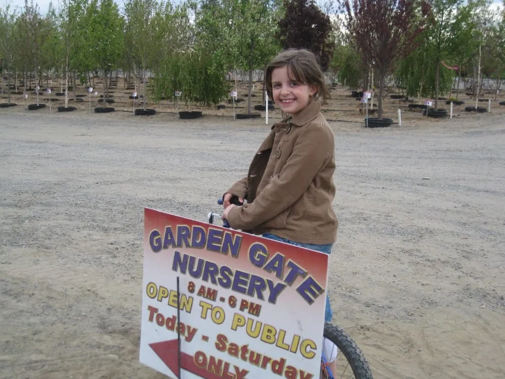 Young girl on a bicycle beside a Garden Gate Nursery Open to Public sign